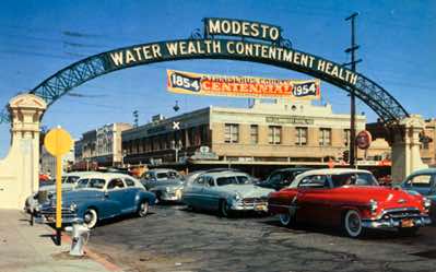 This postcard shows automobiles of mid-20th Century parked on I Street under the arch in 1954. The banner boasts Stanislaus County's Centennial, 1854-1954. The old Claremont Hotel is in the background.
