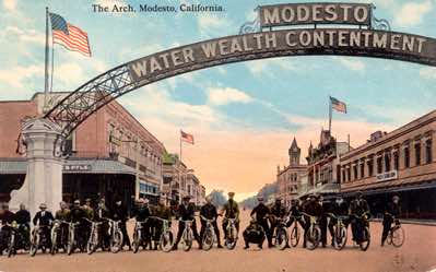 A group of motorcycle enthusiasts, known as the Modesto Motorcycle Club, line up under the arch. There are also a couple of bicycles. Again this is a very early postcard photo, and we note that the welcome sign is gone.